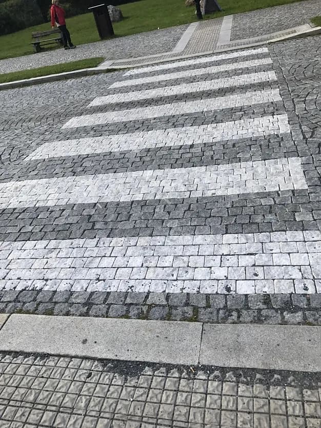 Přechod pro chodce z bílého mramoru a tmavé žulové dlažby v Karlově Studánce, obklopený přírodním kamenem.