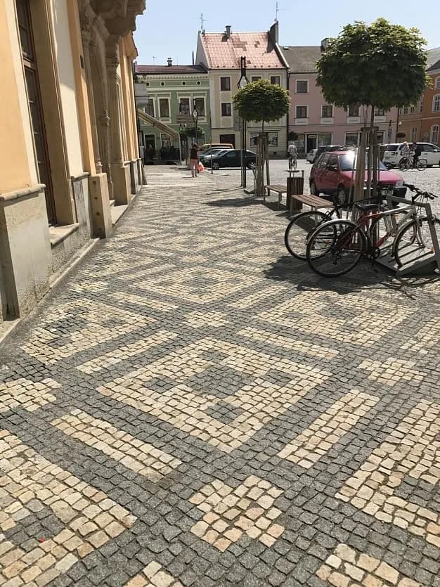 Chodník na náměstí s ornamentální dlažbou z přírodního kamene, lemovaný stromy, lavičkami a stojany na kola.
