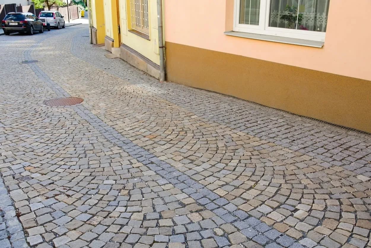 Ulice dlážděná šedožlutými žulovými kostkami v obloukovém vzoru, doplněná kontrastním pásem z menších tmavých kostek.