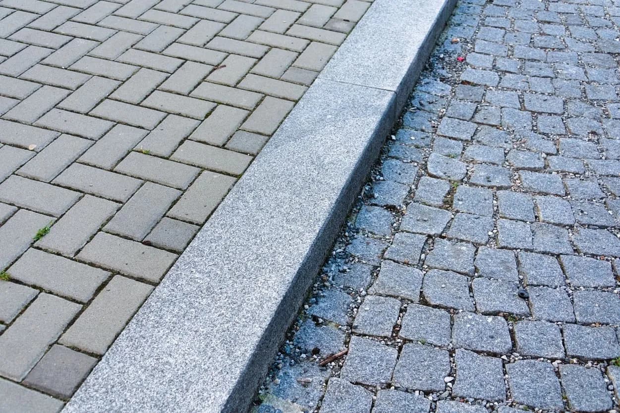 Pohled na přechod mezi chodníkem z betonové dlažby a silnicí s šedými žulovými kostkami, oddělený masivním obrubníkem.