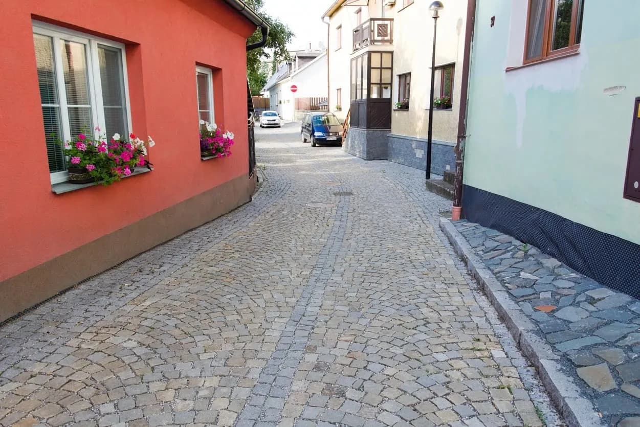 Úzká dlážděná ulička se žulovou dlažbou, lemovaná barevnými domy s květinami v oknech a zvýšeným chodníkem z lomového kamene.