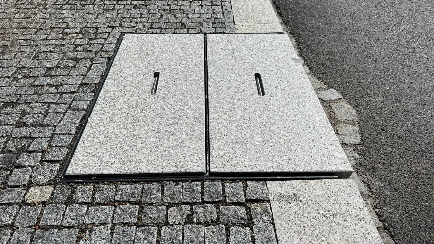Žulový poklop z šedé žuly, zasazený do chodníku z žulových kostek, zajišťuje odolné zakrytí technické infrastruktury.