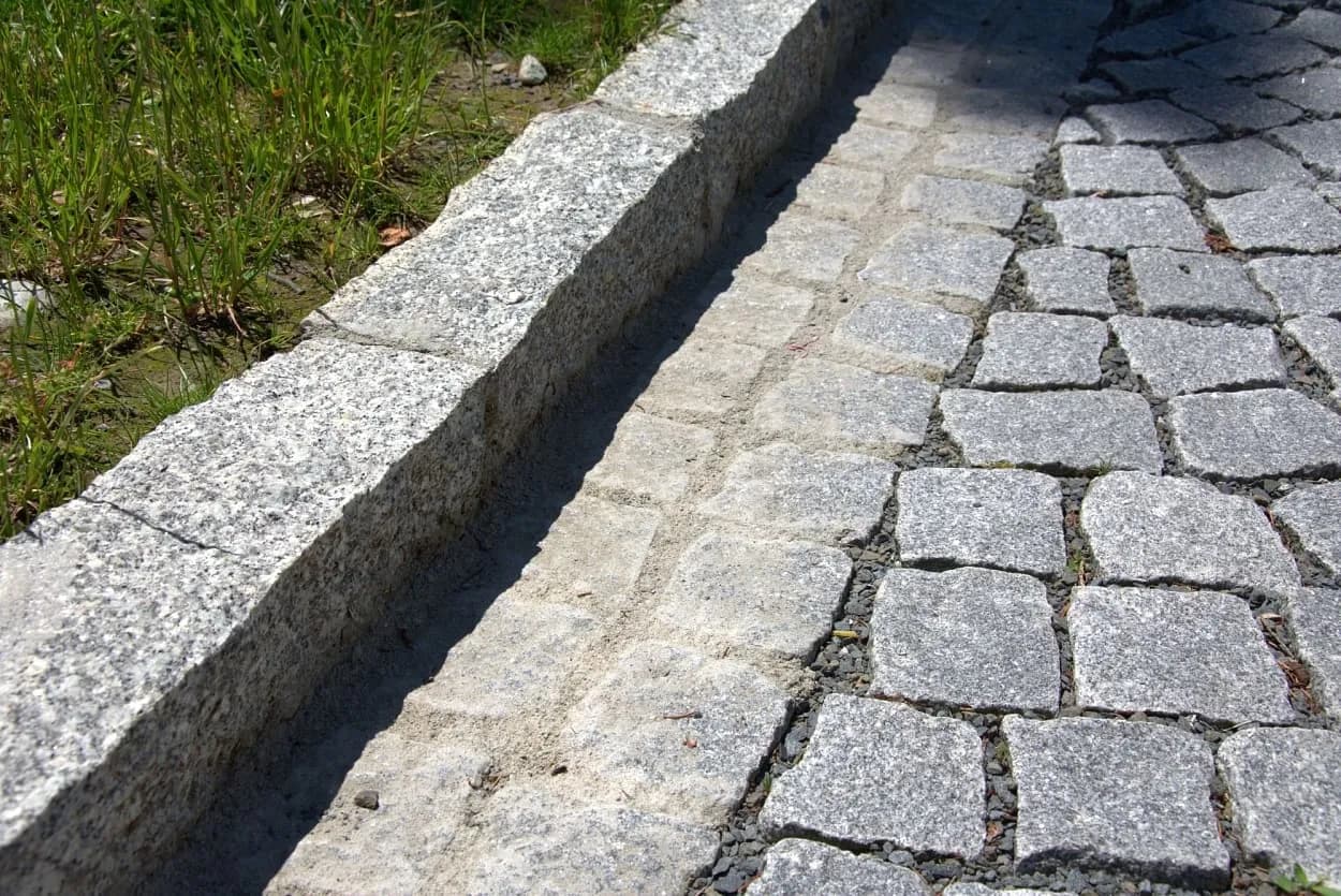 Detail žulového štípaného krajníku se žlábkem a štípanou žulovou dlažbou, která zajišťuje pevnost a odolnost povrchu.